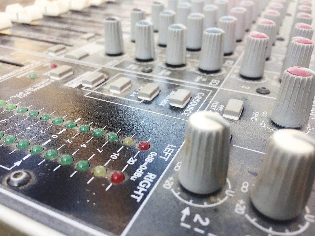 A detailed close-up of an audio mixing console showing numerous control knobs, faders, level meters with LED indicators, and channel strips, depicting specialized audio equipment for mixing.